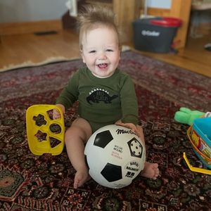 Owen sitting on the floor with a soccer ball and shapes toy, smiling.